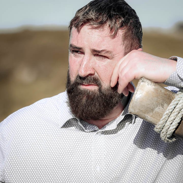 Irish man leaning on a weathered wooden post outdoors, looking off into the distance in natural light.